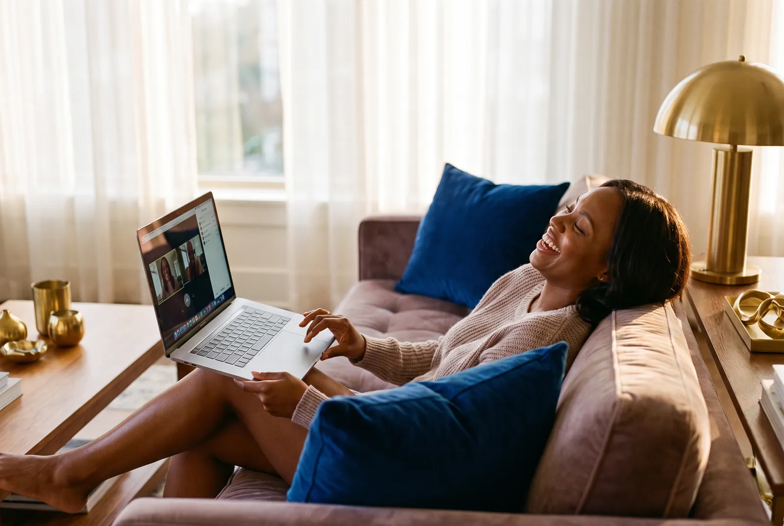 A Black woman relaxed on her sofa at home, laughing during a telehealth video visit on her laptop